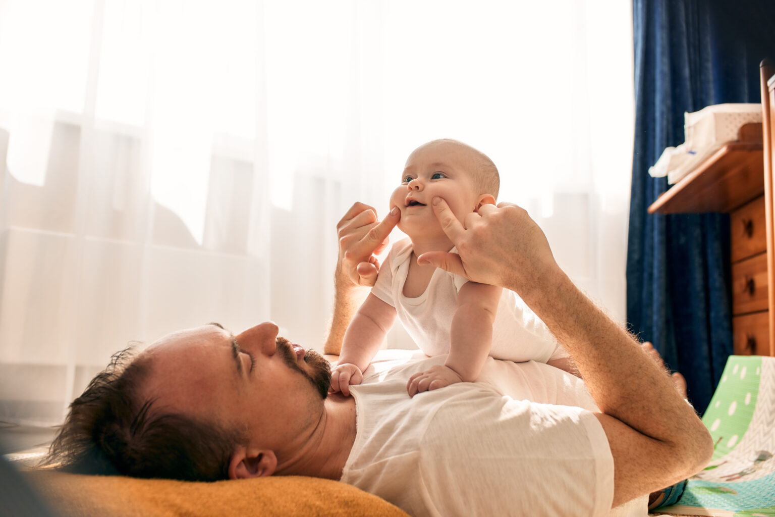 Happy dad holds his newborn baby in his arms while lying on the floor in a bright child's room. The