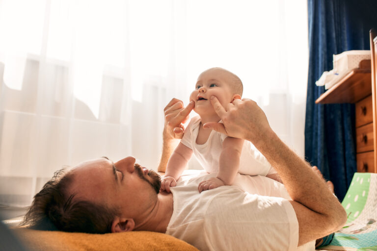 Happy dad holds his newborn baby in his arms while lying on the floor in a bright child's room. The