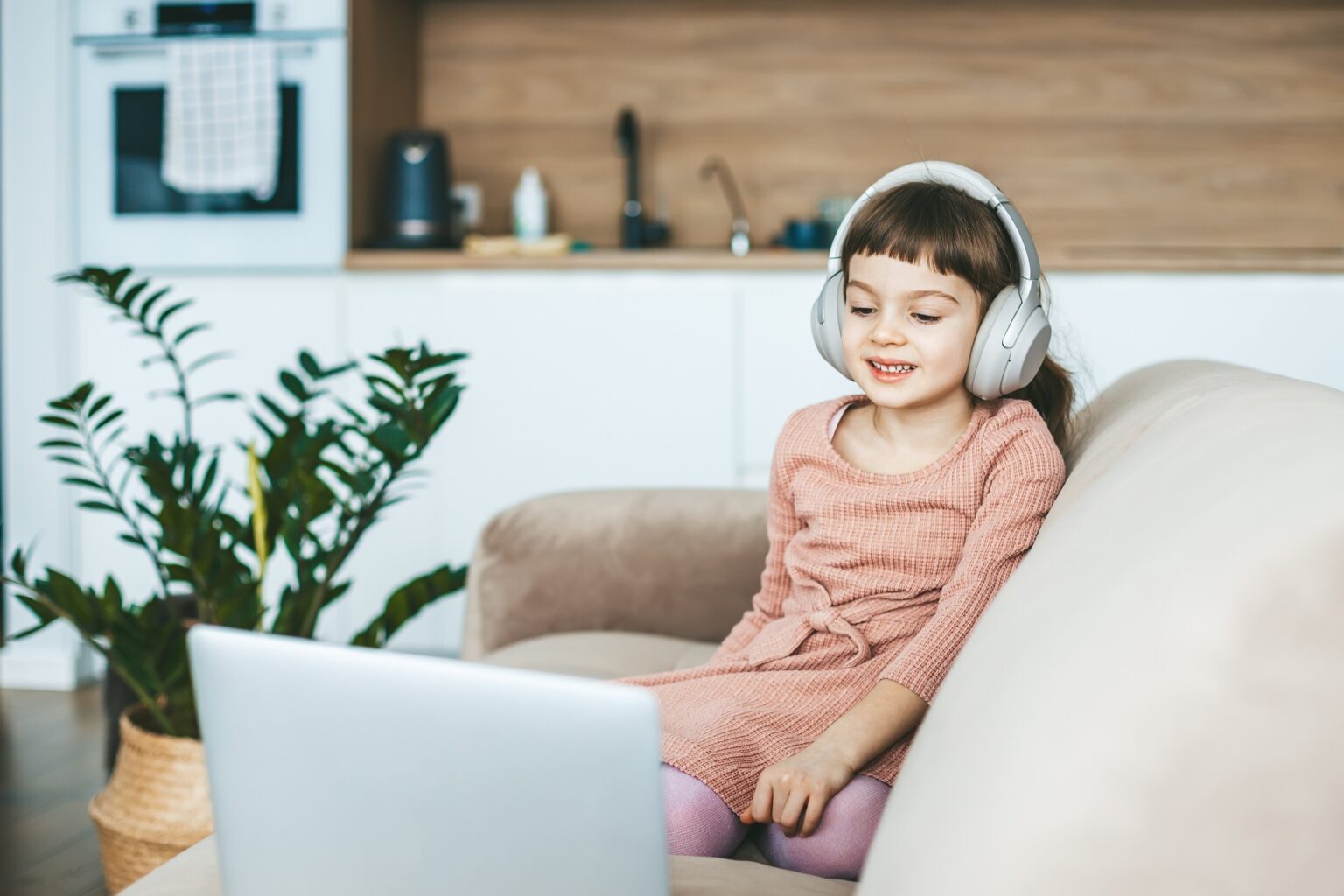A smiling 5-6 years old girl watching a laptop screen, sitting on a beige couch